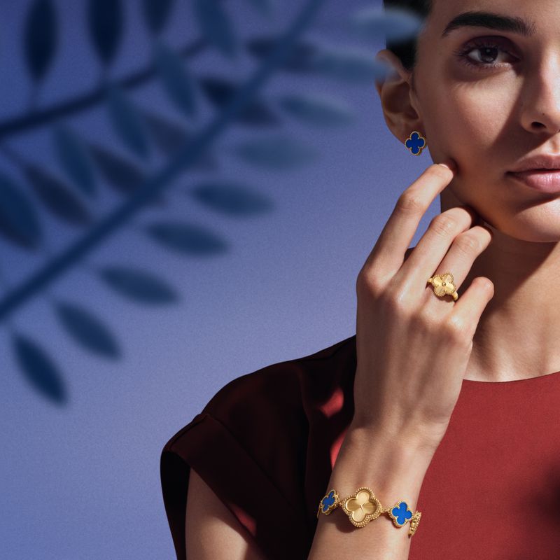 Elegant woman showcasing gold Van Cleef & Arpels jewelry set with clover motifs: earrings, ring, and bracelet against a blue backdrop.