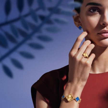 Elegant woman showcasing gold Van Cleef & Arpels jewelry set with clover motifs: earrings, ring, and bracelet against a blue backdrop.