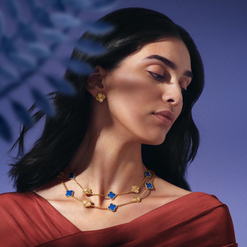 Elegant woman wearing gold earrings and two-strand gold necklace with blue quatrefoil clover designs in a soft violet-blue studio setting.