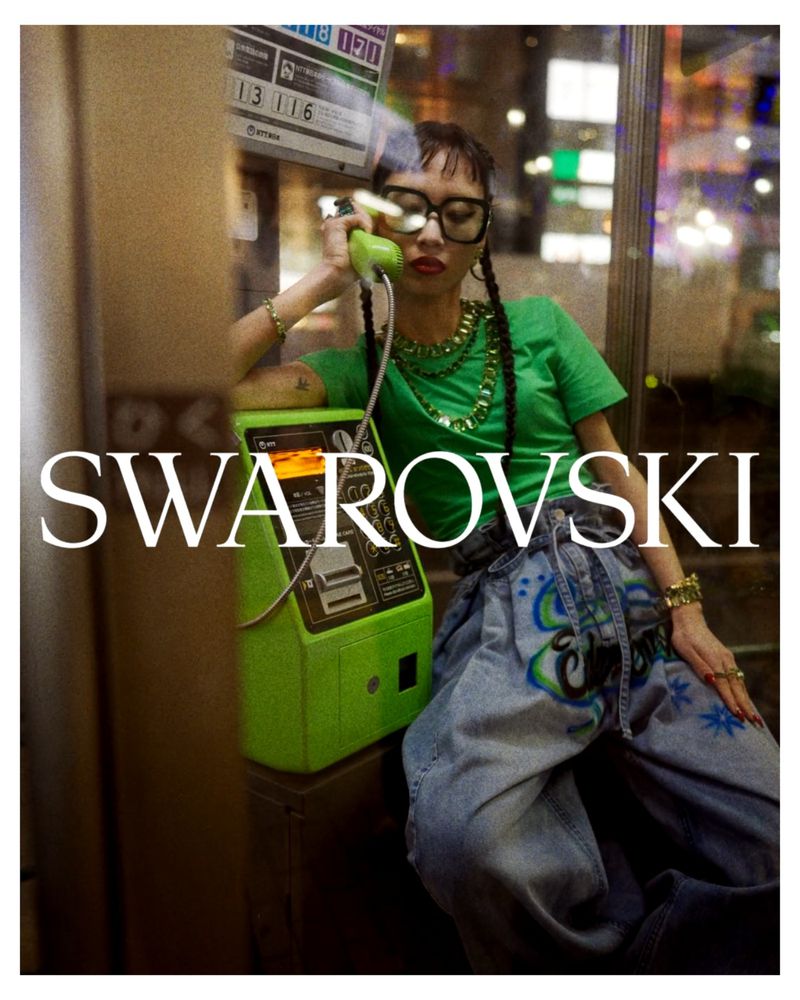 Woman in a green shirt and wide jeans uses a vintage public telephone in a phone booth with the Swarovski logo in the foreground.