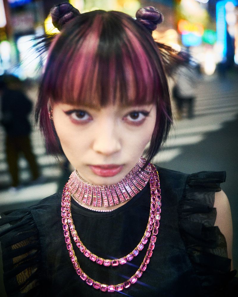 Woman with pink and black hair poses in an urban setting, adorned with layers of pink gem necklaces against blurred lights.