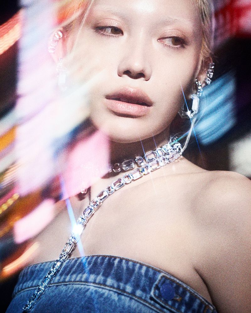 Close-up fashion shot of an adult female with light skin and blonde hair wearing rhinestone jewelry and a denim top against a dark background.
