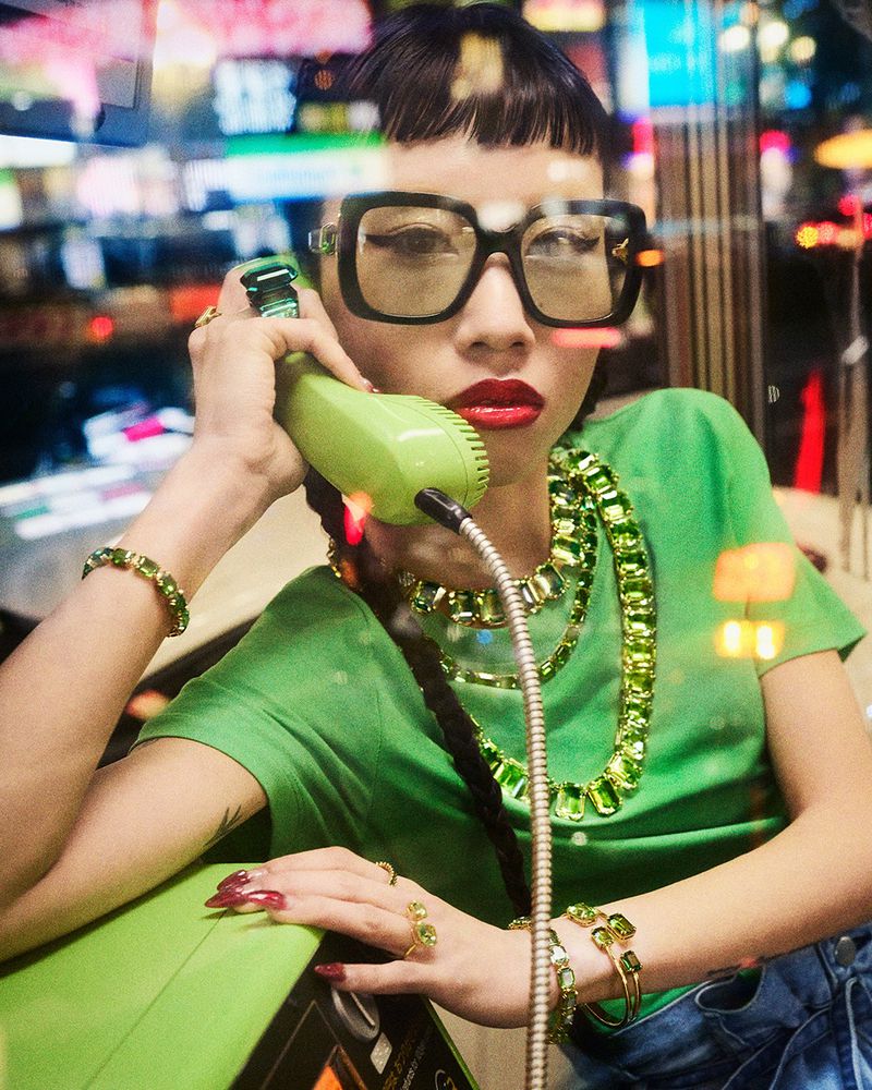 Woman with long dark hair in a green shirt and oversized glasses using a lime green rotary phone in an urban setting at nighttime.