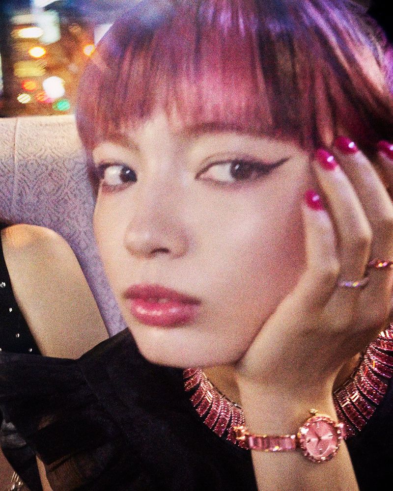 Close-up portrait of an Asian woman with pink-purple hair and manicured nails wearing a pink jeweled watch and necklace in an urban setting.