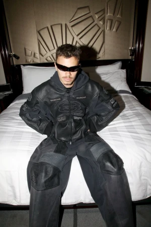 Adult male poses on bed in black jacket and pants. Dark hair, sunglasses, neutral expression, indoor setting, geometric headboard.
