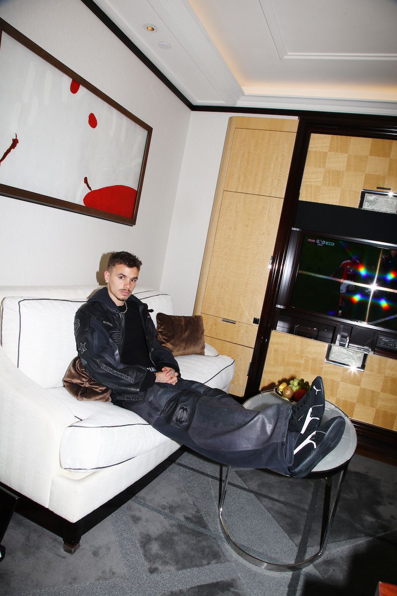 Young adult male lounges on a white sofa in an indoor setting with modern art, wearing a black jacket, top, and oversized grey trousers.