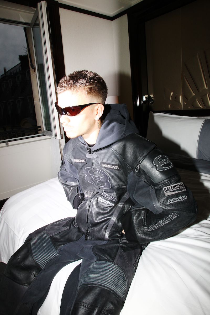 Romeo Beckham wearing a black leather racing jacket and sunglasses, seated on a white hotel bed in an indoor room.