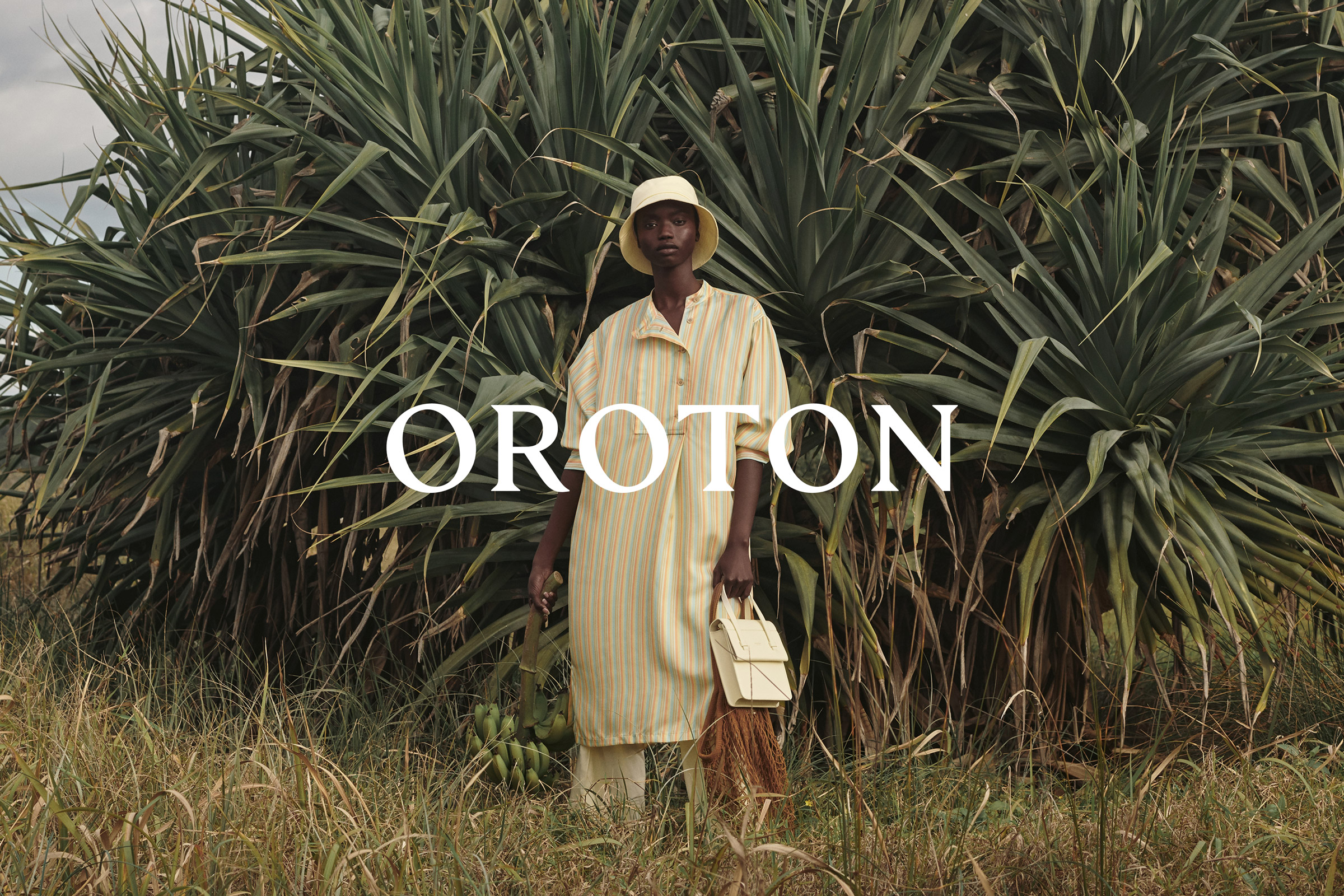 An African-American woman poses in a yellow-striped outfit with a handbag amidst lush greenery. The image features the 'Oroton' logo.