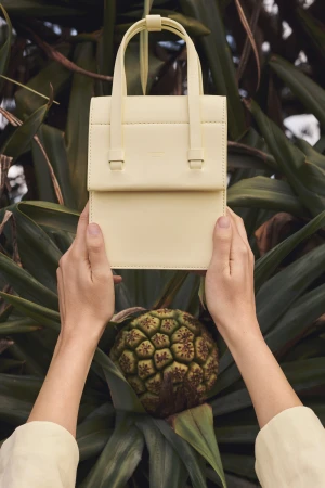 A pale yellow leather handbag with top handle is held up against large green foliage and a tropical fruit in this eye-level shot.