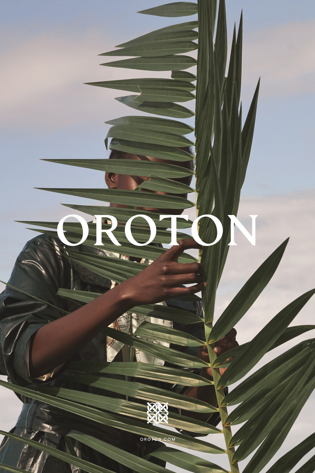 A person poses behind a large green palm leaf against a clear sky in a fashion editorial image. The model is wearing a black top.