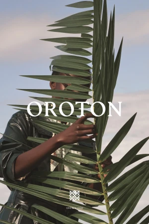 A person poses behind a large green palm leaf against a clear sky in a fashion editorial image. The model is wearing a black top.