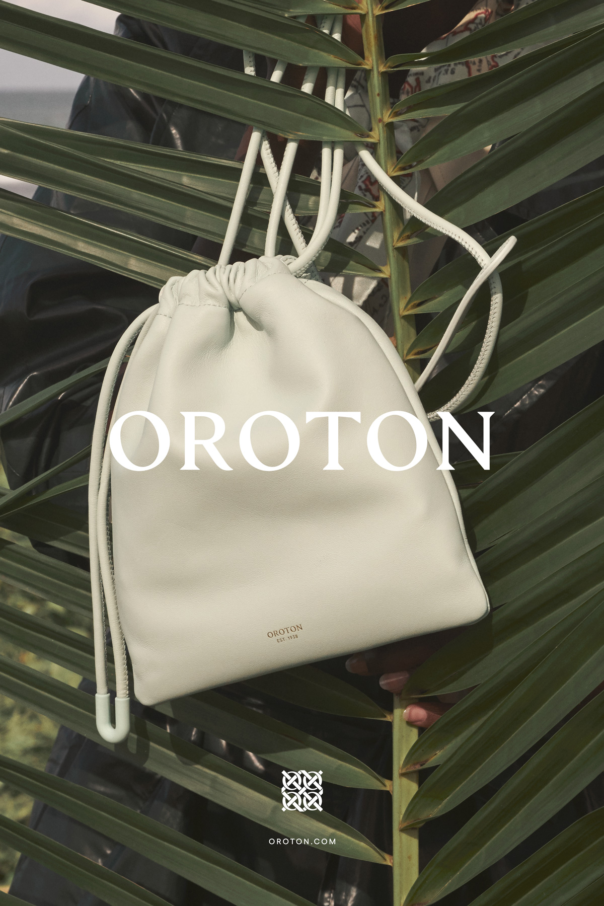 A light-colored leather drawstring handbag is displayed against a background of dark green palm fronds in an eye-level vertical shot.