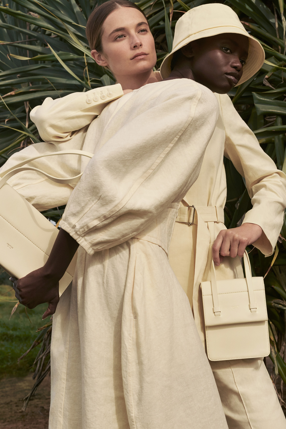 Two female models posing outdoors wearing cream outfits and bucket hats while holding matching handbags for Oroton Spring.