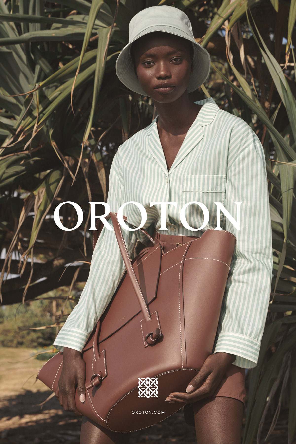 Woman in striped pajamas and bucket hat holds a brown leather tote bag against a green foliage backdrop for Oroton fashion ad.