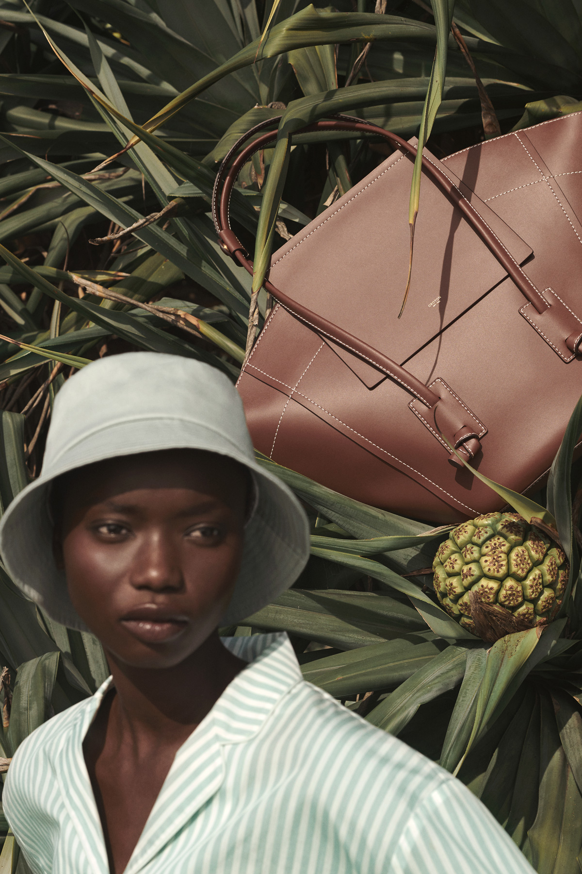 An African woman in a bucket hat and striped shirt poses with a brown leather bag and fruit, surrounded by green tropical foliage.