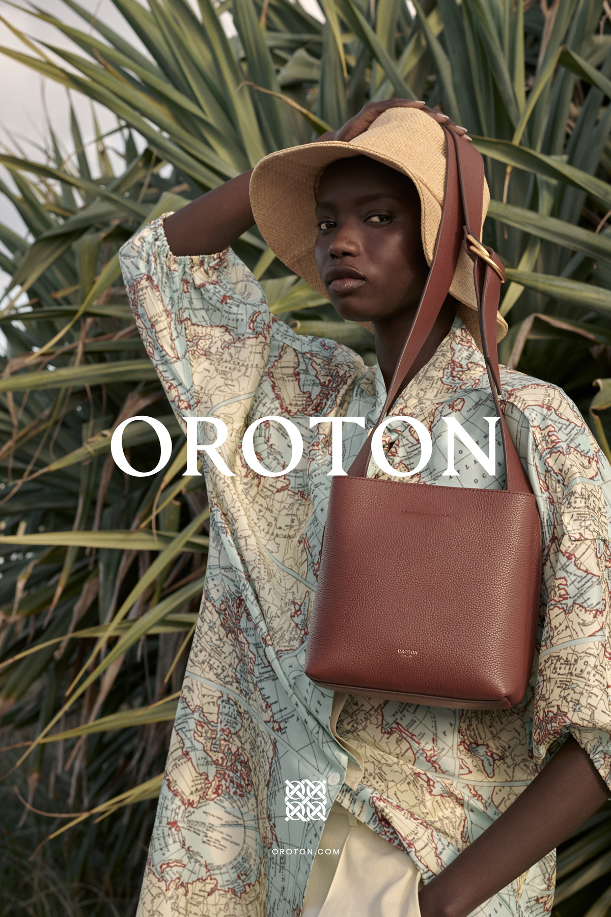 Fashion shot of a Black woman wearing a map-printed shirt and straw sunhat, carrying a brown leather purse against a green foliage background.