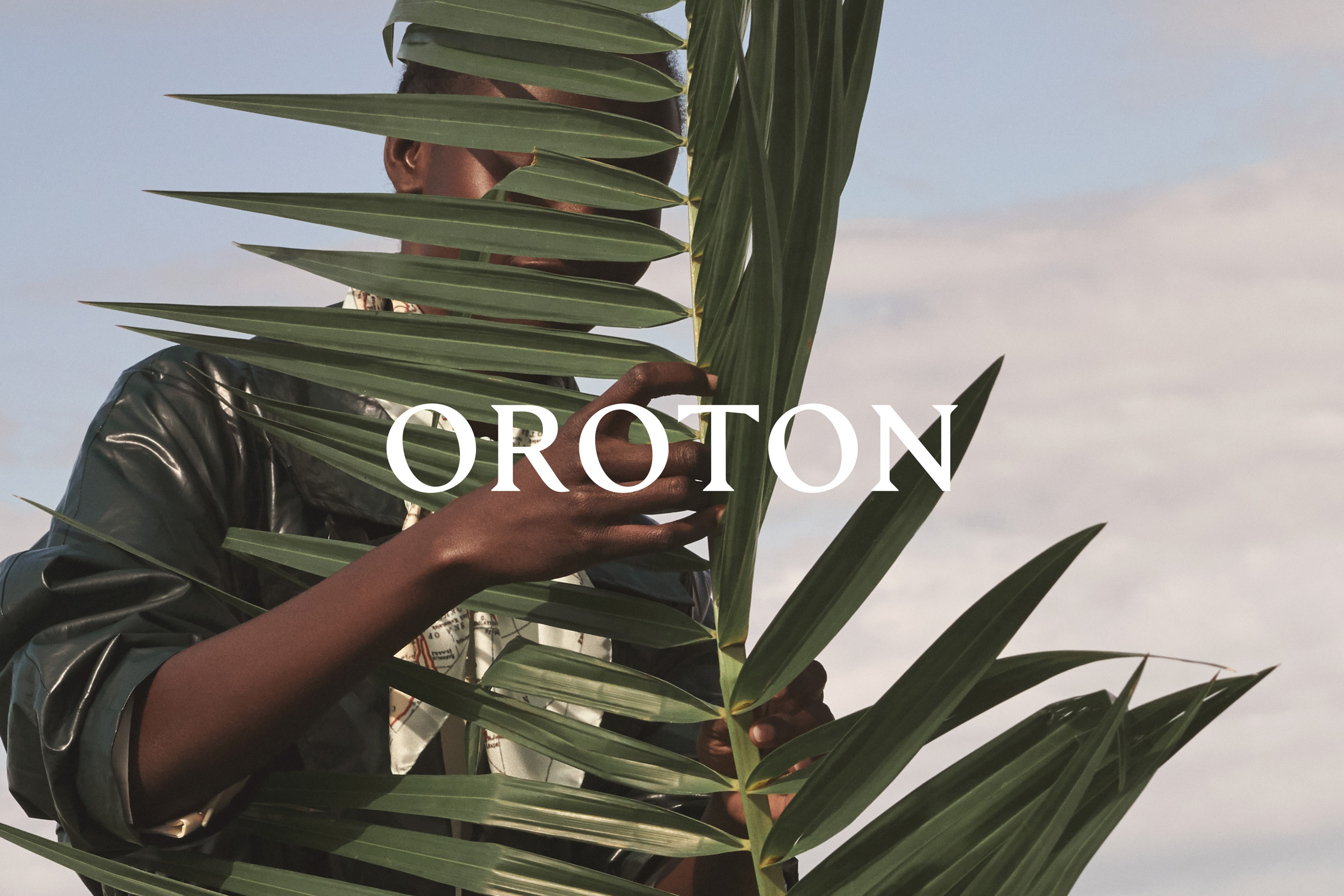 Model presenting Oroton fashion, partially obscured by large green palm leaves. Blue sky backdrop. Dark clothing and white brand text overlay.