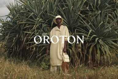 Model wearing striped tunic and bucket hat standing before lush green tropical foliage holding handbag