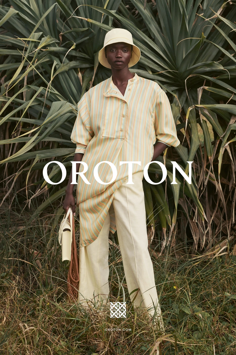 Model wearing a yellow bucket hat, striped tunic, and cream trousers standing among lush tropical foliage.