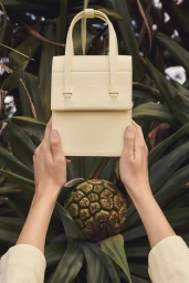 Close-up of hands holding a pale yellow leather handbag against lush green tropical foliage.