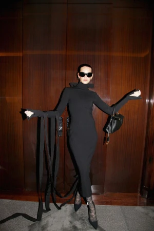 Milena Smit in a black turtleneck dress holding a handbag against dark wood doors for Balenciaga