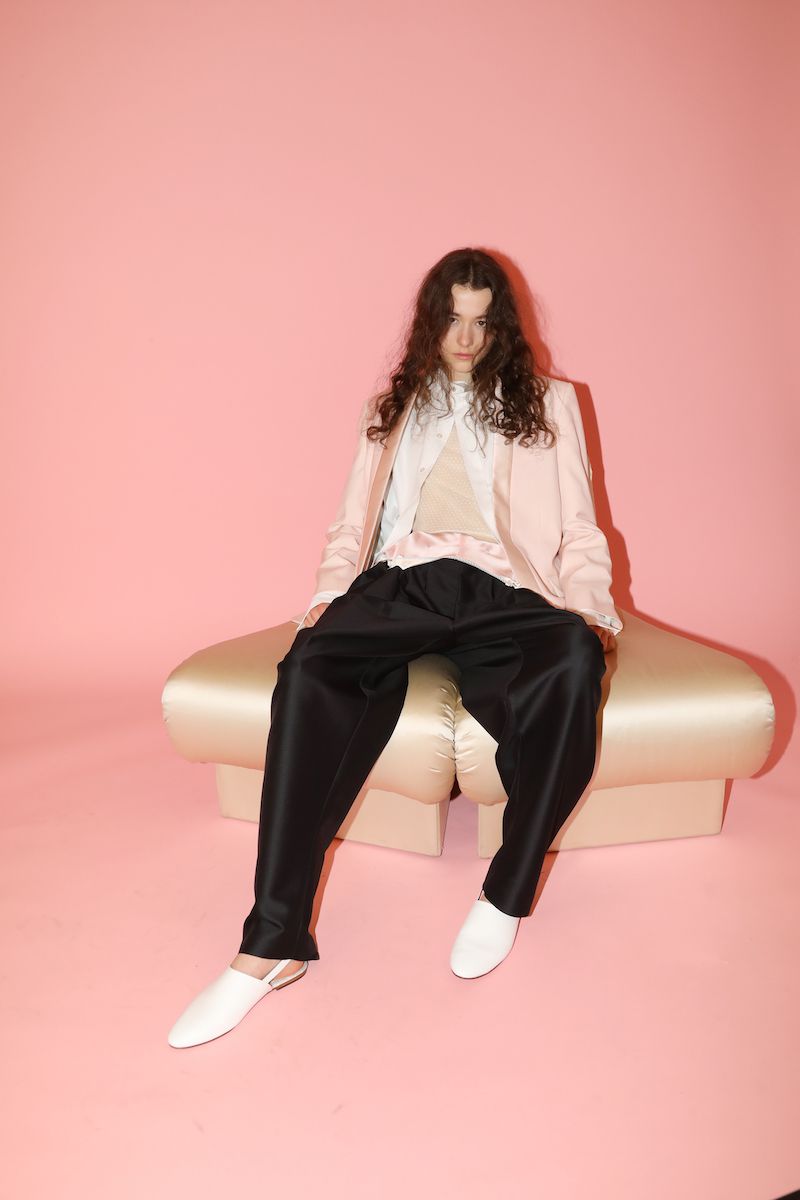 Adult female in a light pink blazer and black trousers is sitting on a beige seat in a photo studio with a pink backdrop.