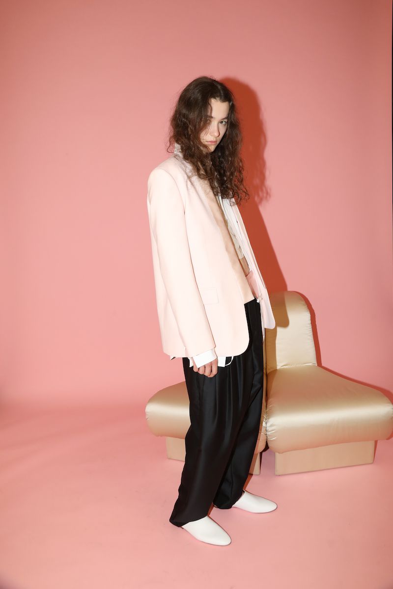 Person in a light pink blazer and black pants poses in a pink studio with a modern, off-white chair. Fashion editorial shot.
