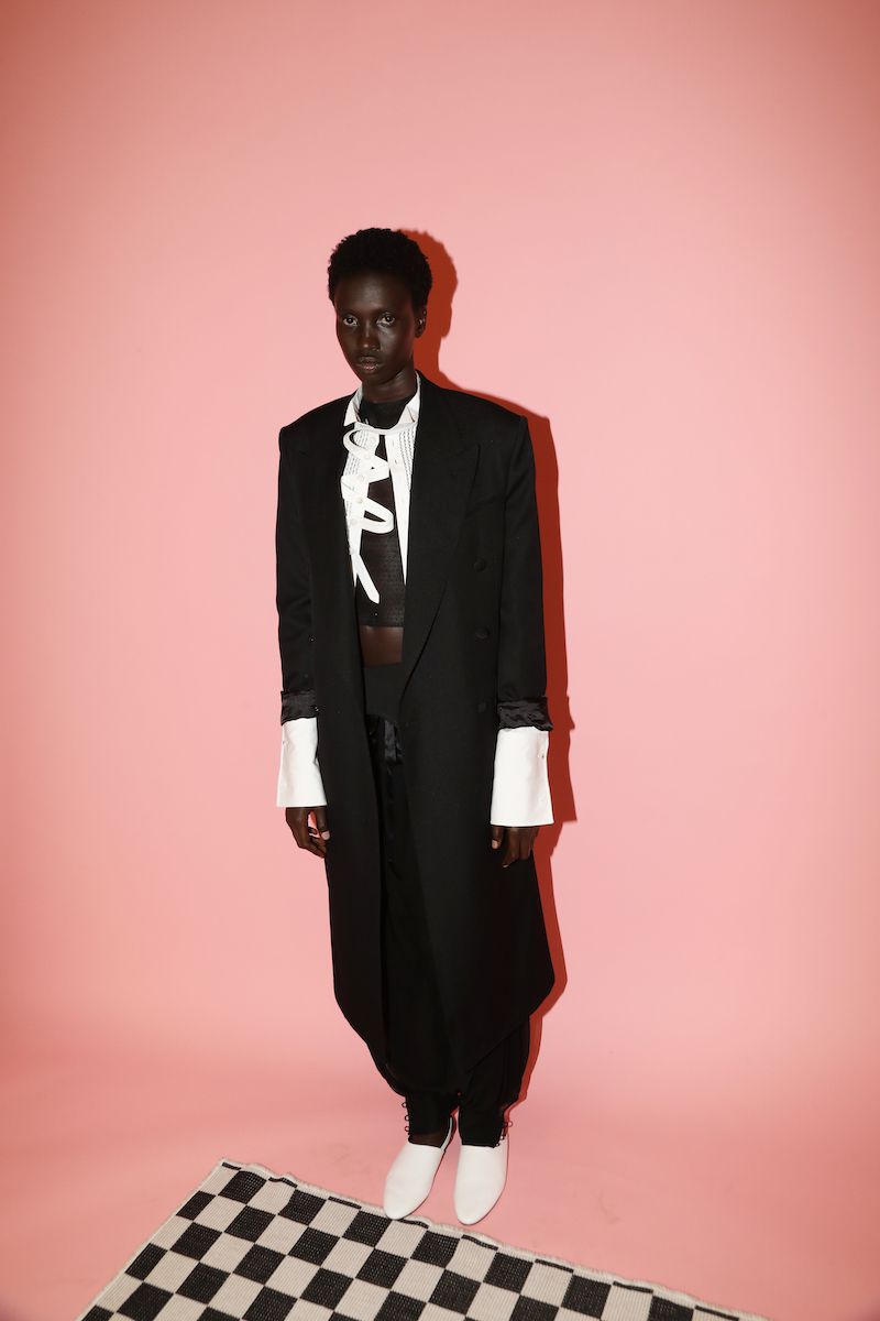 Dark-skinned woman poses in a black overcoat, sheer top, black pants, and white heels, against a pink backdrop and checkerboard rug.