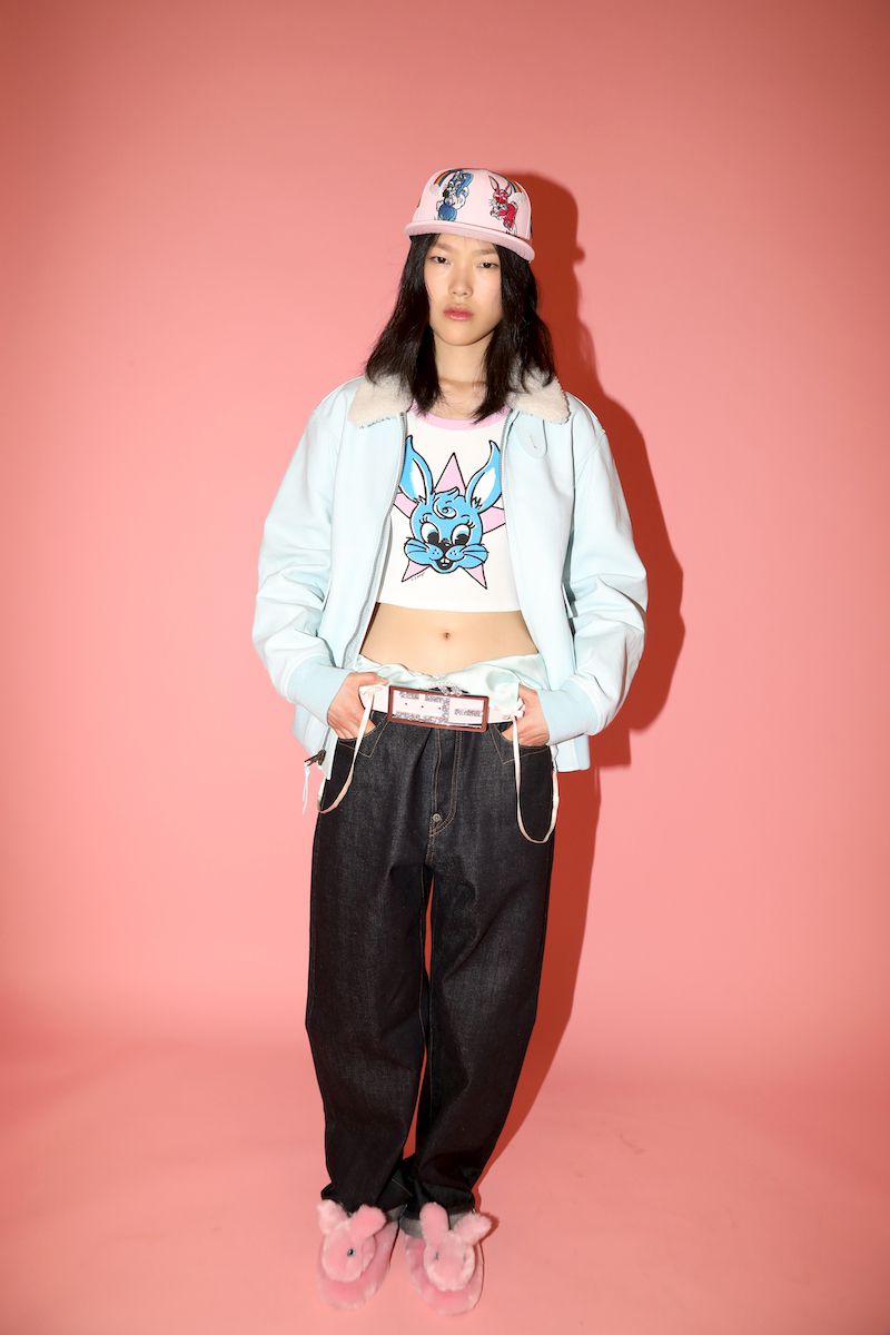 Asian woman in baggy jeans and a cap stands in a studio with a pink background, showcasing casual fashion with a playful and youthful vibe.