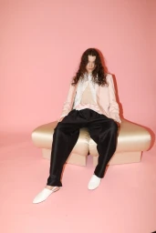 Model with long dark hair seated on a pale ottoman wearing a light pink blazer and black trousers against a pink backdrop