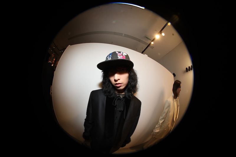 Portrait of a person with long dark hair, wearing a black suit jacket and hat, posing in front of a white wall. Fisheye lens effect.