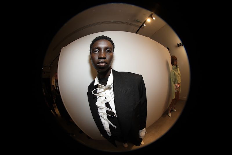 Close-up, low-angle fisheye lens shot of an African male fashion model in a black suit, white shirt and tie, posing indoors in a photo studio.