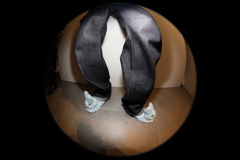 A pair of blue jeans with the legs bent at an angle, worn with blue bunny slippers, set against a neutral background.