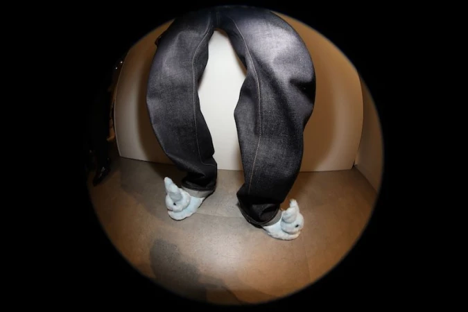 A pair of blue jeans with the legs bent at an angle, worn with blue bunny slippers, set against a neutral background.