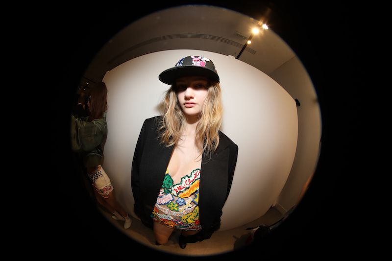 Wide-angle shot of a woman in a black blazer and patterned dress, posing indoors with a neutral expression and wearing a baseball cap.