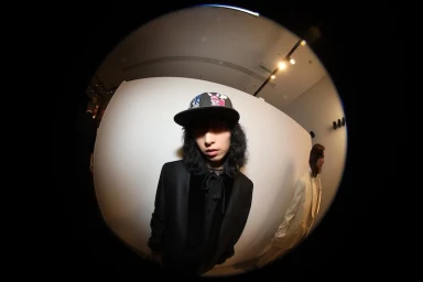 Fisheye portrait of a person with long dark hair wearing a patterned baseball cap and black blazer against a white studio wall.