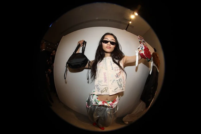 Fish-eye lens fashion shot of a woman in sunglasses holding a black handbag and floral accessories against a plain white backdrop.