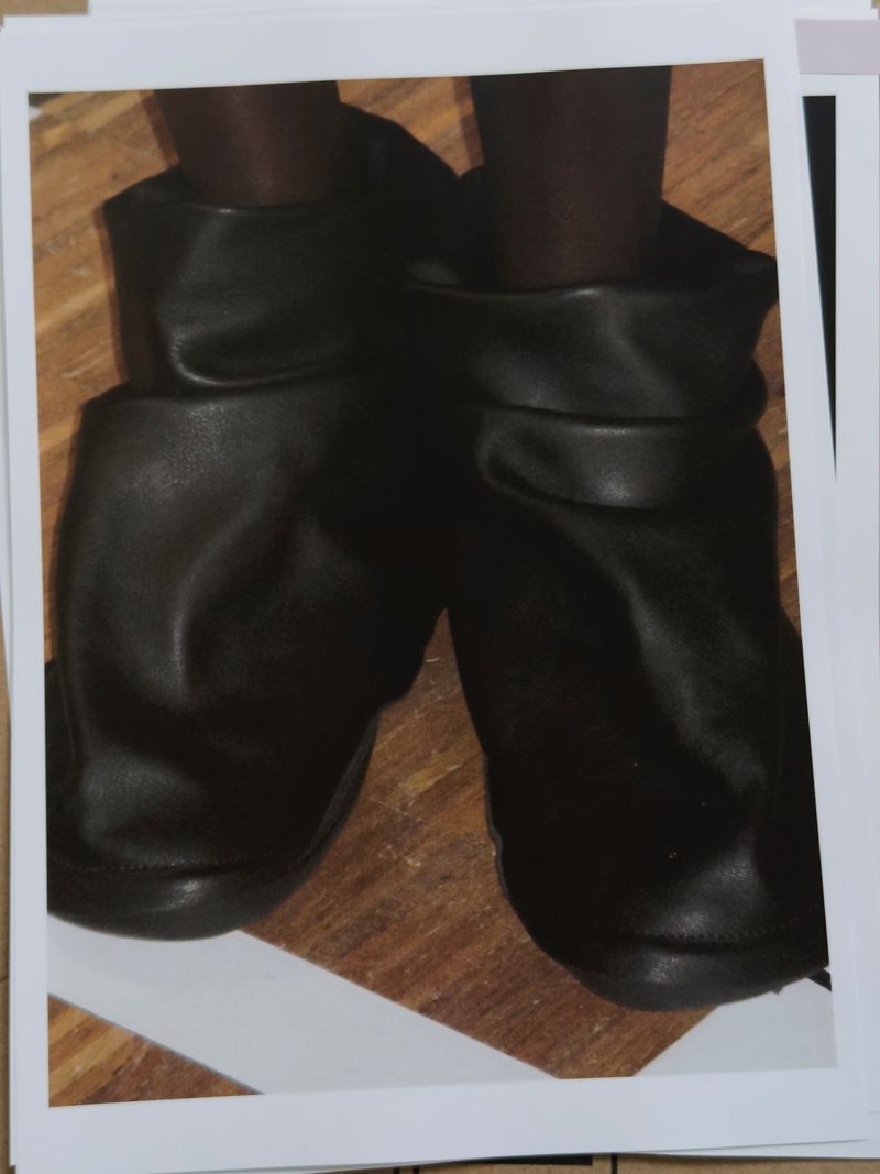 Close-up studio shot of black leather booties with brown leggings on a wood background, presented on white cards with a light grey border.