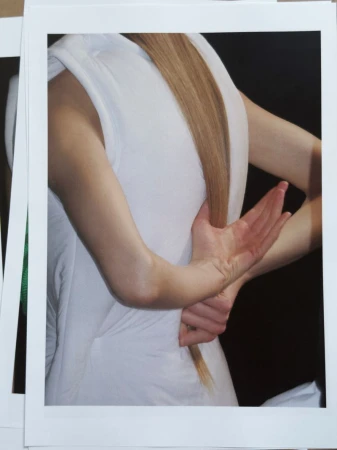 Woman with light brown hair and a white dress is shown from behind. The woman has her arms crossed behind her back.