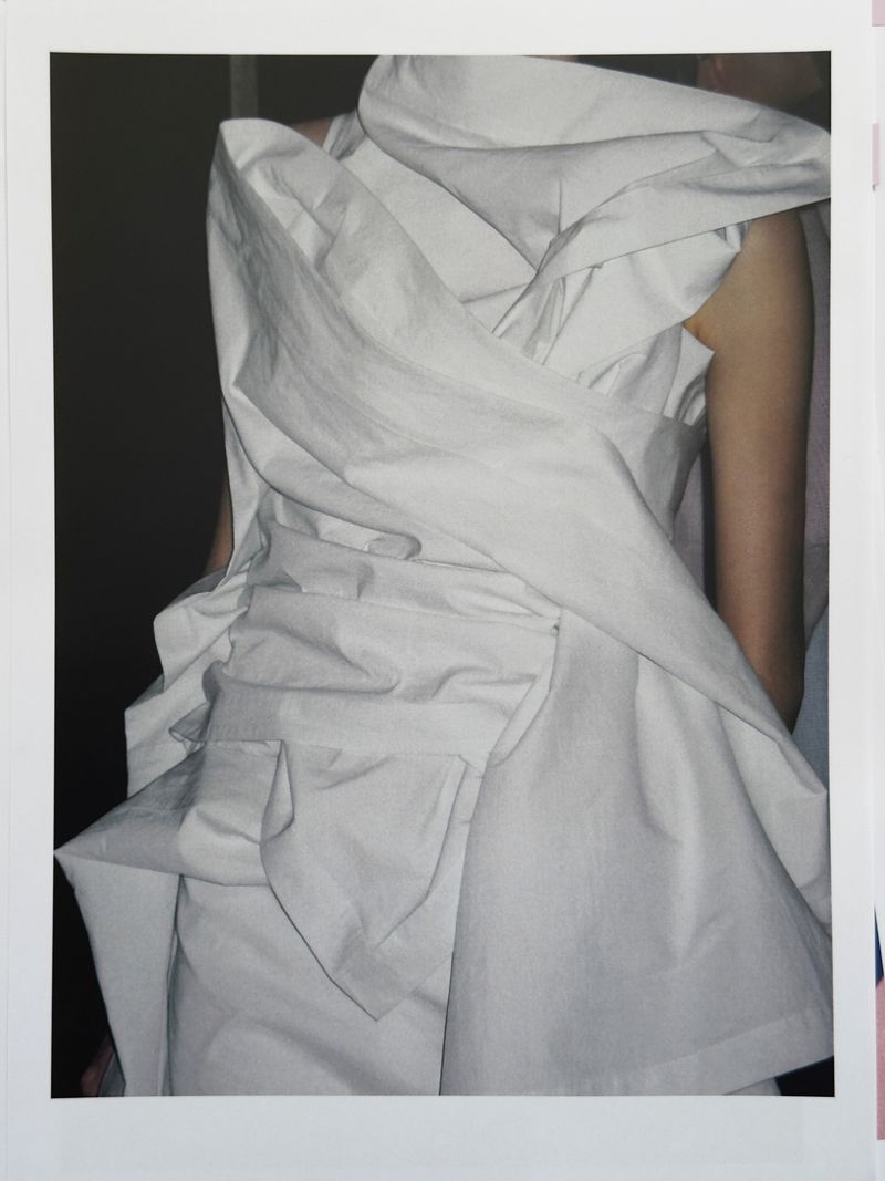Fashion shot featuring a person wearing an elaborate white dress with draping fabric, set against a muted dark background in a studio-style setting.