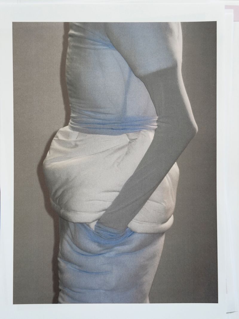 Monochromatic art photograph features a person draped in light blue and white fabric, with an arm bent across their body. 