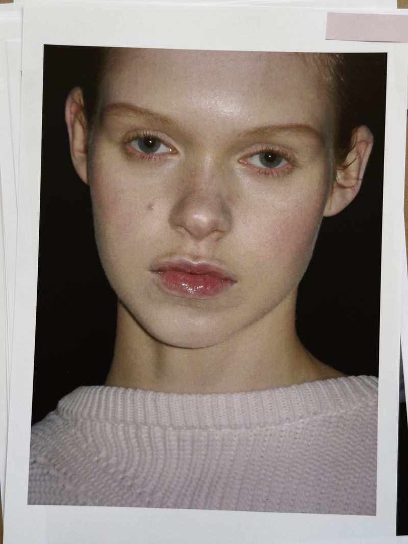 Close-up portrait of a fair-skinned young woman with light hair and blue eyes, wearing a pink sweater, photographed on photo prints.