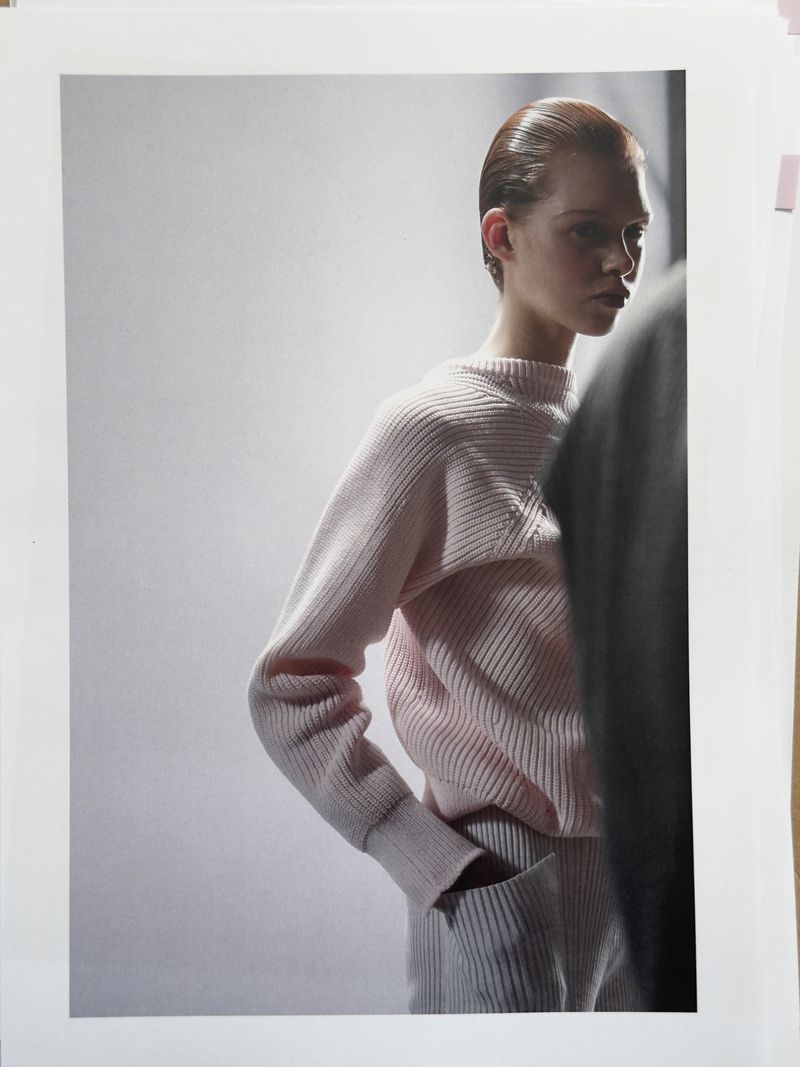 Fashion shot of a young woman with slicked-back hair, wearing a pink ribbed sweater and pants, hands in pockets, against a white backdrop.