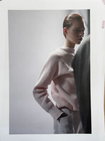 Fashion shot of a young woman with slicked-back hair, wearing a pink ribbed sweater and pants, hands in pockets, against a white backdrop.