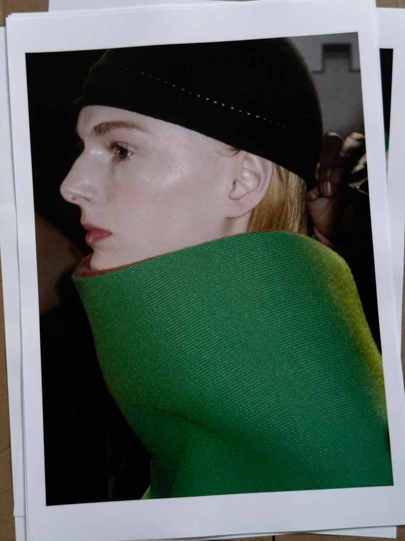 Profile portrait of a fair-skinned person wearing a black beanie and vibrant green shawl, looking left against a dark background indoors.