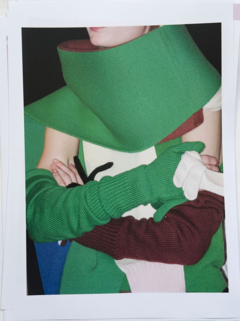 Close-up studio shot of a person wearing a large green and maroon collar and green sleeves over white gloves, against a dark backdrop.