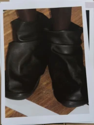 Close-up of a person wearing black slouchy leather boots standing on a wooden floor, featured in the Issey Miyake x Byredo project.