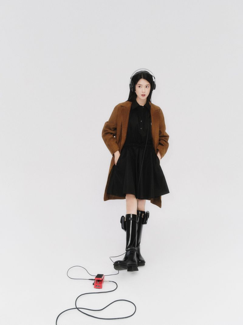Full shot of a young adult woman wearing a brown coat and black dress with headphones in a white studio with a red guitar pedal.