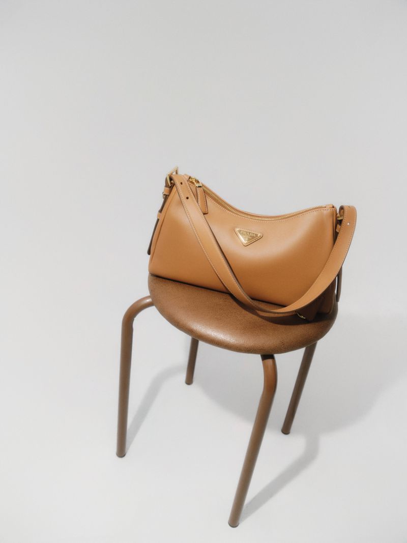 Tan leather Prada handbag with gold logo on a brown stool against a white backdrop in a studio setting for a fashion editorial.