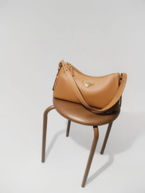 Tan leather Prada handbag with gold logo on a brown stool against a white backdrop in a studio setting for a fashion editorial.